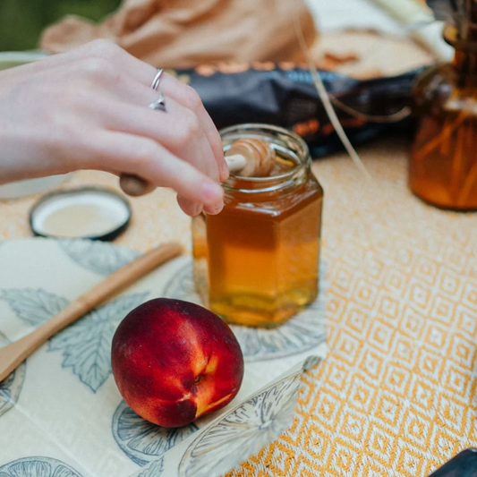 a woman's hand dipping a honey stirrer into a jar of raw, unpasteurised local cheshire heather honey set on a yellow picnic blanket