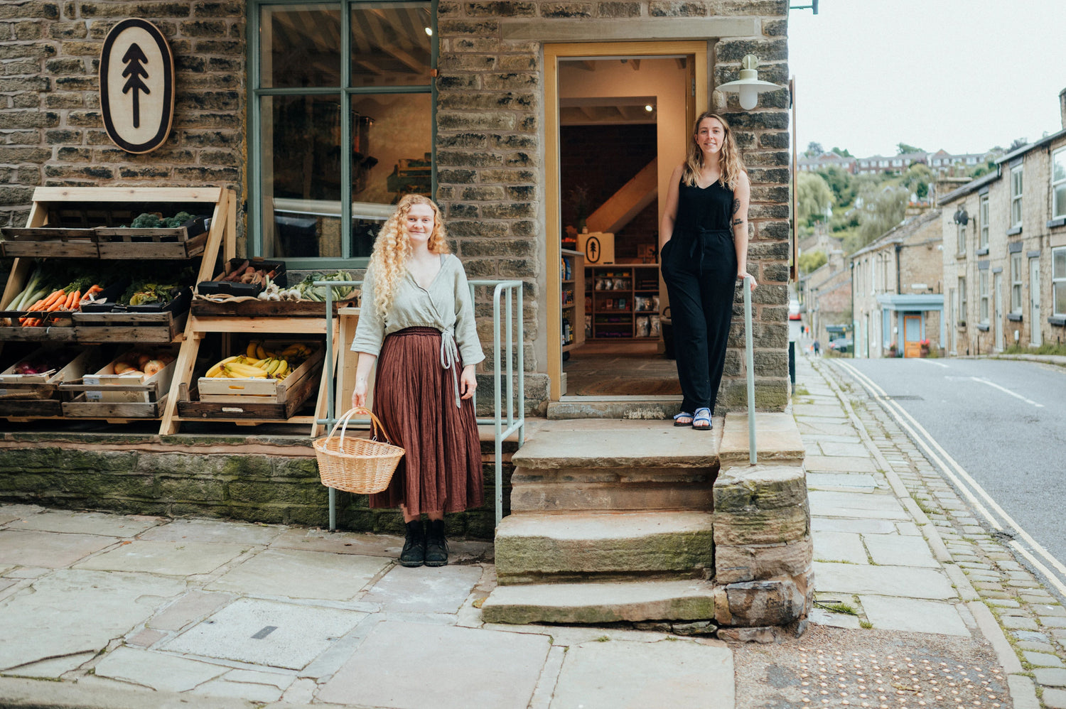 two women stood outside of their organic grocer, refill shop holding baskets and smiling at the camera, there is fresh vegetable stand stood to their right