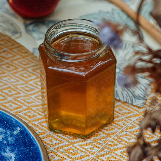 a jar of raw, unpasteurised local cheshire heather honey set on a yellow picnic blanket with blurred flowers in the foreground