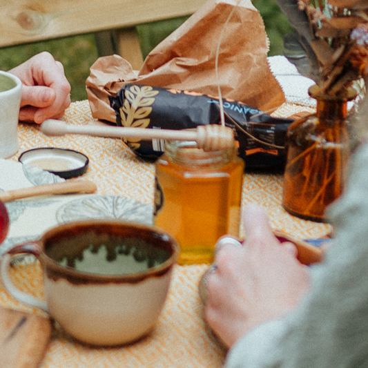 a honey stirrer set on top of a jar of raw, unpasteurised local cheshire heather honey set on a yellow picnic blanket wamongst flowers, cups of tea and two women chatting